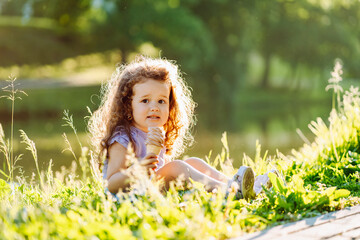 Little curly smiling girl eating chocolate ice cream while sitting on the grass in the summer park.
