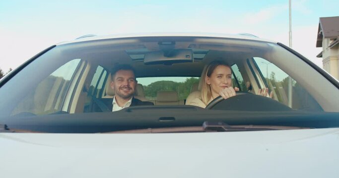 Young attractive couple driving in auto. Returning from work to home, talking to each other and laughing. Outdoors. Front view.