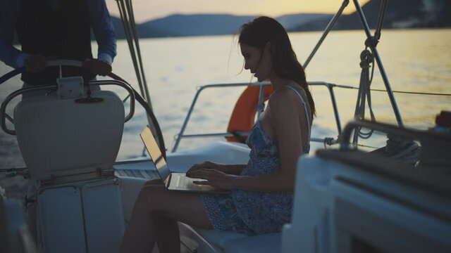Man and woman sailing on calm water in evening. Brunette typing on laptop. Summer vacation at sea, sailing and travelling concept.