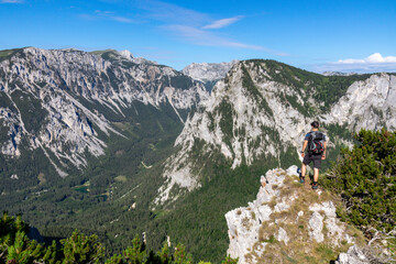 A man with a hiking backpack standing on the rock with a view on the Alpine mountain chains in Austria, Hochschwab region. The slopes are partially overgrown with small bushes, higher parts baren.