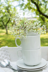 A pyramid of white cups and saucers, a branch of white flowers, two teaspoons on a linen napkin with curly on a blurred background of a summer garden. Still life of preparation for summer tea.