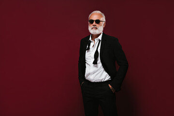 Man in suit and white shirt wears sunglasses. Grey haired guy with beard in classic outfit posing on isolated background..