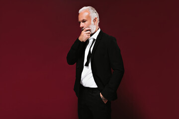Adult man dressed in suit thoughtfully poses on burgundy background. Pensive bearded guy with grey...
