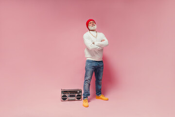 Serious man in jeans and white sweater posing on pink background with record player. Bearded guy...