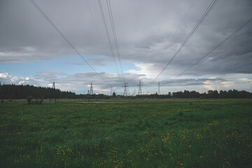 electric transit lines through meadows and agricultural fields. Meadow with green grass in front, river, electric pylons and forest trees in background. Cloudy sky in spring evening