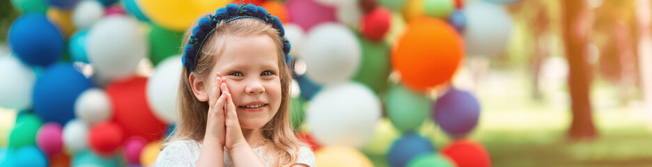 Banner. A beautiful, little girl, of European appearance demonstrates emotions of joy and surprise. A sweet girl folds her hands near her cheek and smiles against the background of colorful balloons