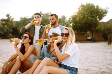 Group of friends cheers and drink beers on the beach. Young  friends sitting together sitting near the sea and enjoy summer party. Beach holiday and summer vacation concept.