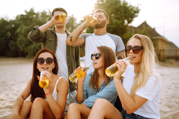 Group of friends cheers and drink beers on the beach. Young  friends sitting together sitting near the sea and enjoy summer party. Beach holiday and summer vacation concept.