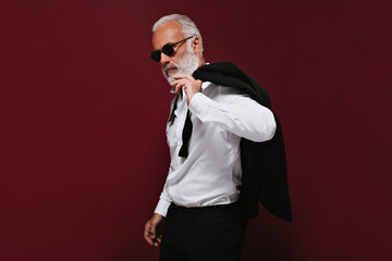 Adult man in white shirt and black sunglasses poses on burgundy background. Charming guy with grey beard in glasses and fashionable suit posing..