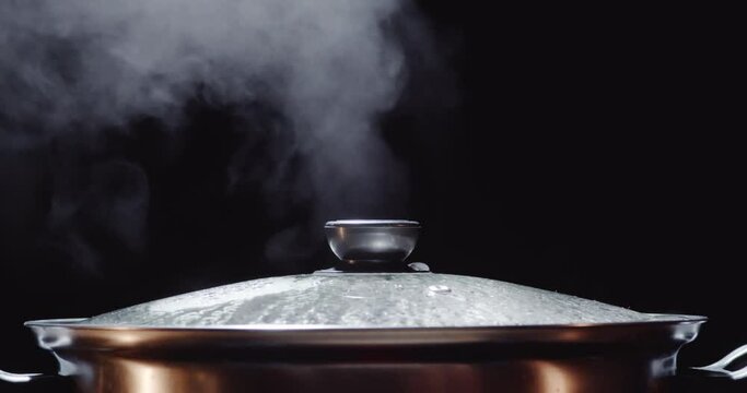 Slow motion of hot pot or shabu-shabu pot made of stainless steel with a glass lid, water boiling, hot steam rising on a black screen, orange lights reflecting inside, a hand reached to open the lid.