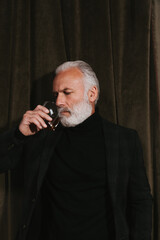 Adult man in black outfit drinking whiskey. Pensive guy with grey beard in dark shirt and jacket posing on velvet background..