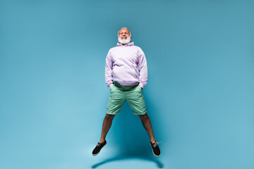 Fototapeta na wymiar White haired man in stylish outfit jumps on blue background. Bearded adult guy in lilac hoodie and green shirt posing on isolated backdrop..