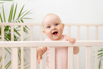 a smiling baby girl in a crib in a pink bodysuit for six months stands holding on to the side and laughs