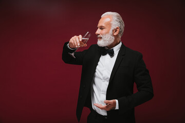 Bearded man in suit drinks champagne. Smiling grey haired adult guy in white shirt posing with wine glass on burgundy background..