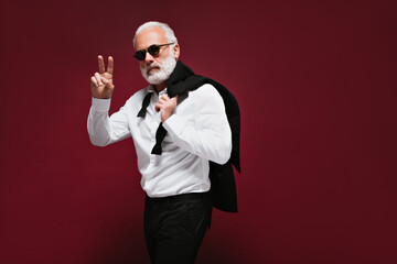 Positive man in suit showing peace sign on burgundy background. Stylish guy with white beard in...