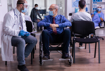 Handicapped senior man in wheelchair talking with doctor wearing protection mask sitting in waiting area of new normal clinic. Specialist medic explaning treatment during coronavirus global pandemic