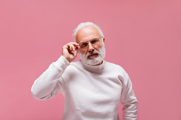 Serious man puts on eyeglasses and poses on pink background. Stylish grey haired guy with beard in...