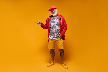 Fototapeta premium Man in red jacket and orange shorts pointing to place for text. White bearded guy with tattoos in modern bright clothes smiling..