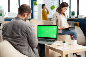 Over the sholder of manager sitting on couch holding laptop with green screen, mockup desktop while diverse team working on background. Multiethnic people planning project on chroma key display