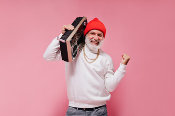 Man in red hat posing with record player on pink background. Joyful adult guy with grey beard in...