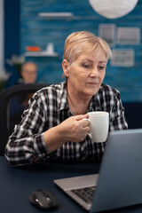 Fototapeta premium Senior entrepreneur woman reading on computer enjoying a cup of coffee and husband and reading a book. Elderly woman in home living room using moder technoloy laptop for communication sitting at desk