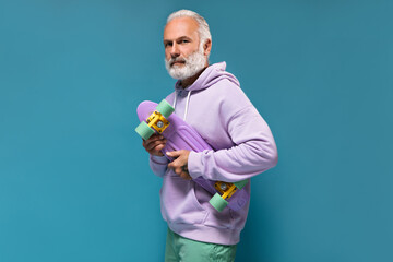 Stylish man in hoodie looks into camera and holds longboard on blue background. Fashionable guy...