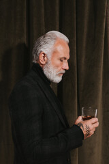 Handsome man in black jacket holds whiskey glass. Thoughtful guy with white hair in dark outfit...