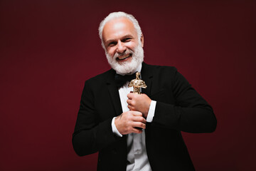 Cool man in black suit laughing and holding Oscar statuette. Smiling guy with grey beard and...
