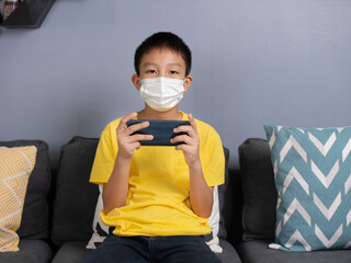 Happy asian boy wearing face masks sit on sofa at home holding phone looking at screen waving hand video calling distance friend online in mobile.