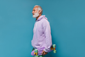 Adult man in street style outfit holding longboard on blue background. White haired guy in lilac...
