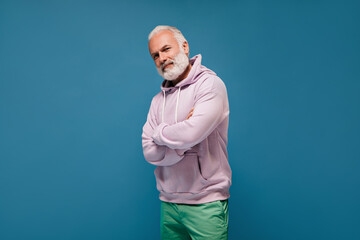 Fototapeta premium Portrait of man in sport style hoodie on blue background. Cool adult guy with grey hair in lilac sweatshirt and green pants posing..
