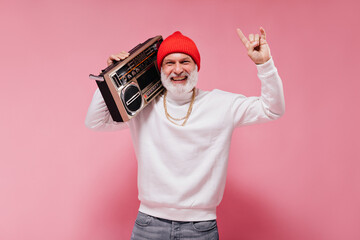 Positive man in red hat happily posing with record player. Joyful guy in white sweatshirt, orange...