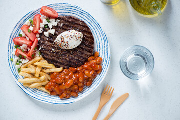 Grilled balkan pljeskavica with kaymak cheese, red tomatoes, fries and prebranac, flatlay over white stone background, horizontal shot