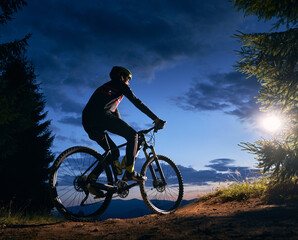 Rear view of cyclist riding bike under blue evening sky with clouds. Silhouette of male bicyclist riding bicycle on the trail in night mountain forest. Concept of sport, biking and active leisure.