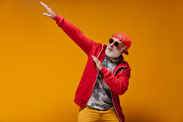 White bearded man in sunglasses and cap posing on orange background. Cool guy in red hoodie and...