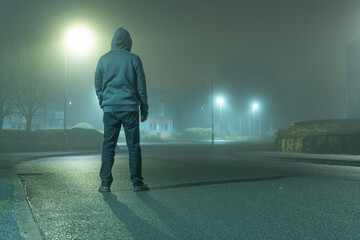 A mysterious hooded figure with back to camera, standing on a road in a light industrial urban area. On a moody, foogy, winters night - Powered by Adobe