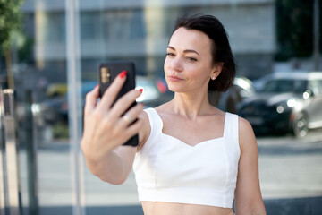 stylish adult woman in white takes a selfie portrait on the phone while standing in the middle of an urban street against a background of glass