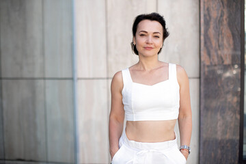 an adult business woman in a white suit holds her hands in her pockets, looks at the camera and smiles while standing on urban street