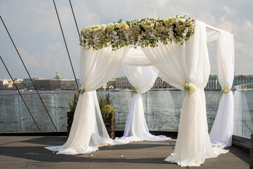 Beautiful wedding arch decorated with white rose flowers installed on the river embankment in the city. Wedding ceremony in sunny summer day.
