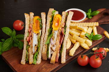 Club-sandwich with french fries. Sandwich with bacon, fried egg. Grilled and pressed toast with bacon, fried egg, tomato and lettuce served on wooden cutting board.