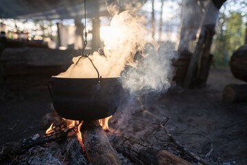 Bowler with escaping steam hangs over a campfire, in which food is prepared, at sunset, against the...