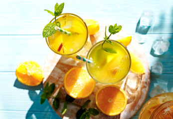 Glasses of lemonade with oranges, ice cubes and mint leaves on blue wooden background. Top view of lemonade. Summer cold drinks under direct sun shines. Caustic reflection effect.