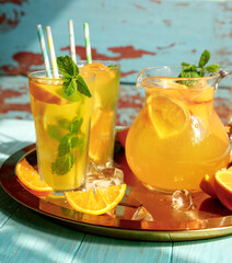 Glasses of lemonade with lemons, oranges and mint leaves on blue wooden background. Summer cold drinks under direct sun shines.