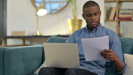 Young African Man with Documents working on Laptop on Sofa 