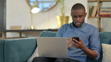 Young African Man working on Smartphone and Laptop on Sofa 