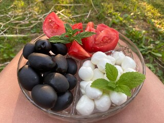 Portrait of a plate with large black olives lying on a glass plate next to red tomatoes and white mozzarella balls decorated with green mint and lemon balm leaves. The plate is on the lap of the photo