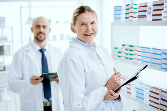 pharmacist with a clipboard and her colleague standing near a pharmacy display case .