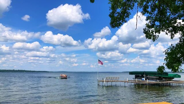 Beautiful Minnesota lake scene as a pontoon slowly approaches shore. A hand held clip with with an American flag waving over the boat dock, close tree branches, and fluffy white clouds under blue sky.