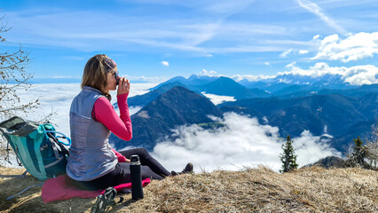 A woman in hiking outfit sipping a tea from thermos with the panoramic view from the top of Alpine...