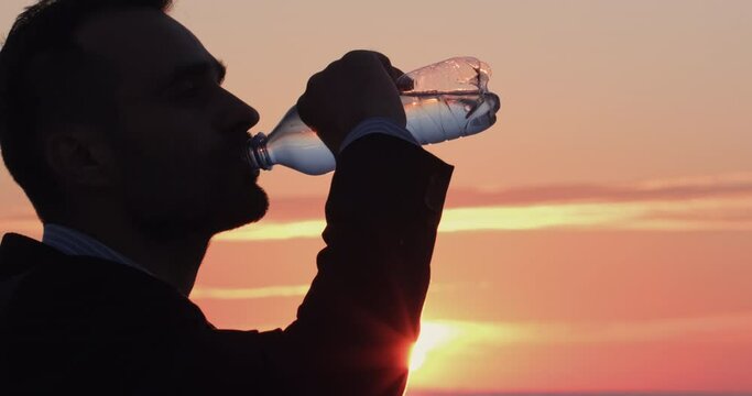 A portrait of a man in silhouette who quenches his thirst by drinking water from a bottle at dawn. Water is the main valuable mineral of the planet.